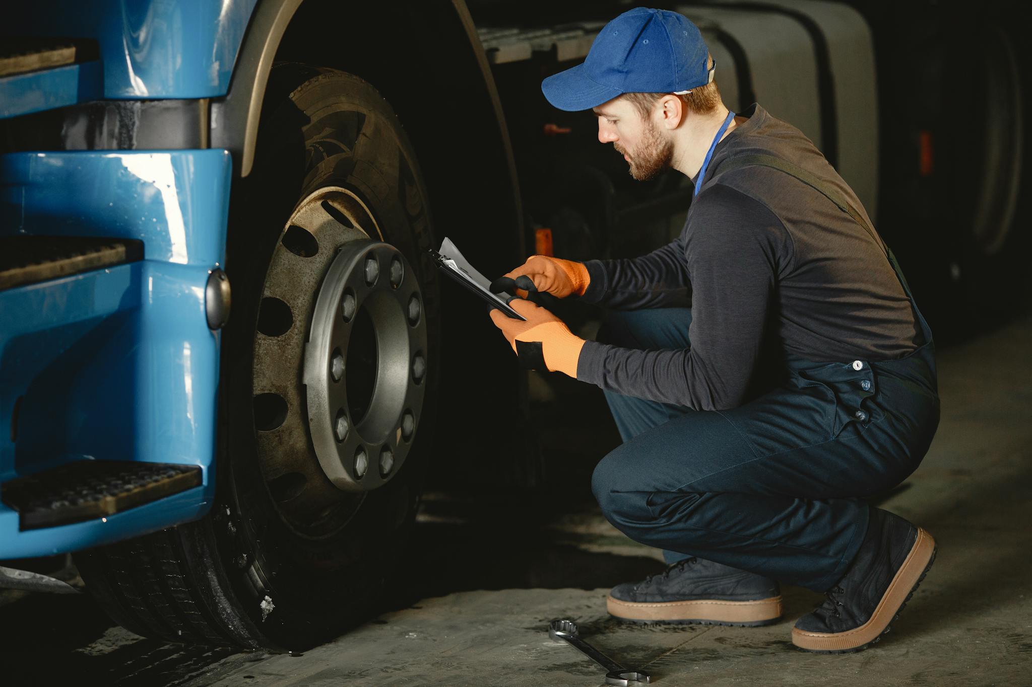 Man in Blue Cap and Working Uniform Sitting Beside Truck Wheel and Reading a Paper